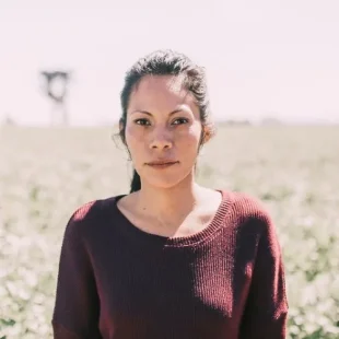 Confident woman in maroon sweater stands in thriving agricultural field under bright blue sky.