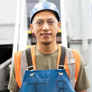 Young man in safety gear confidently stands in industrial workshop environment.